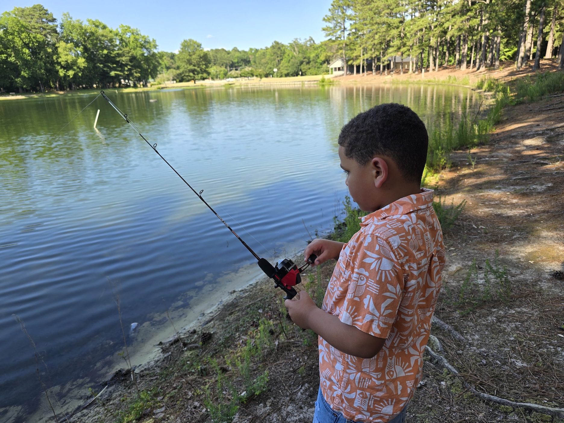 A child in an orange shirt fishes with a black kids fishing pole with a red spincast reel.