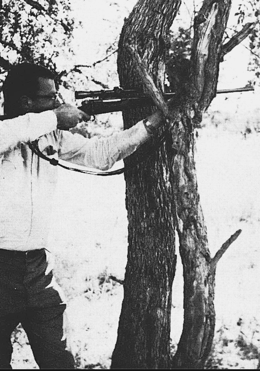 A black and white photo of a man resting a rifle in a tree.