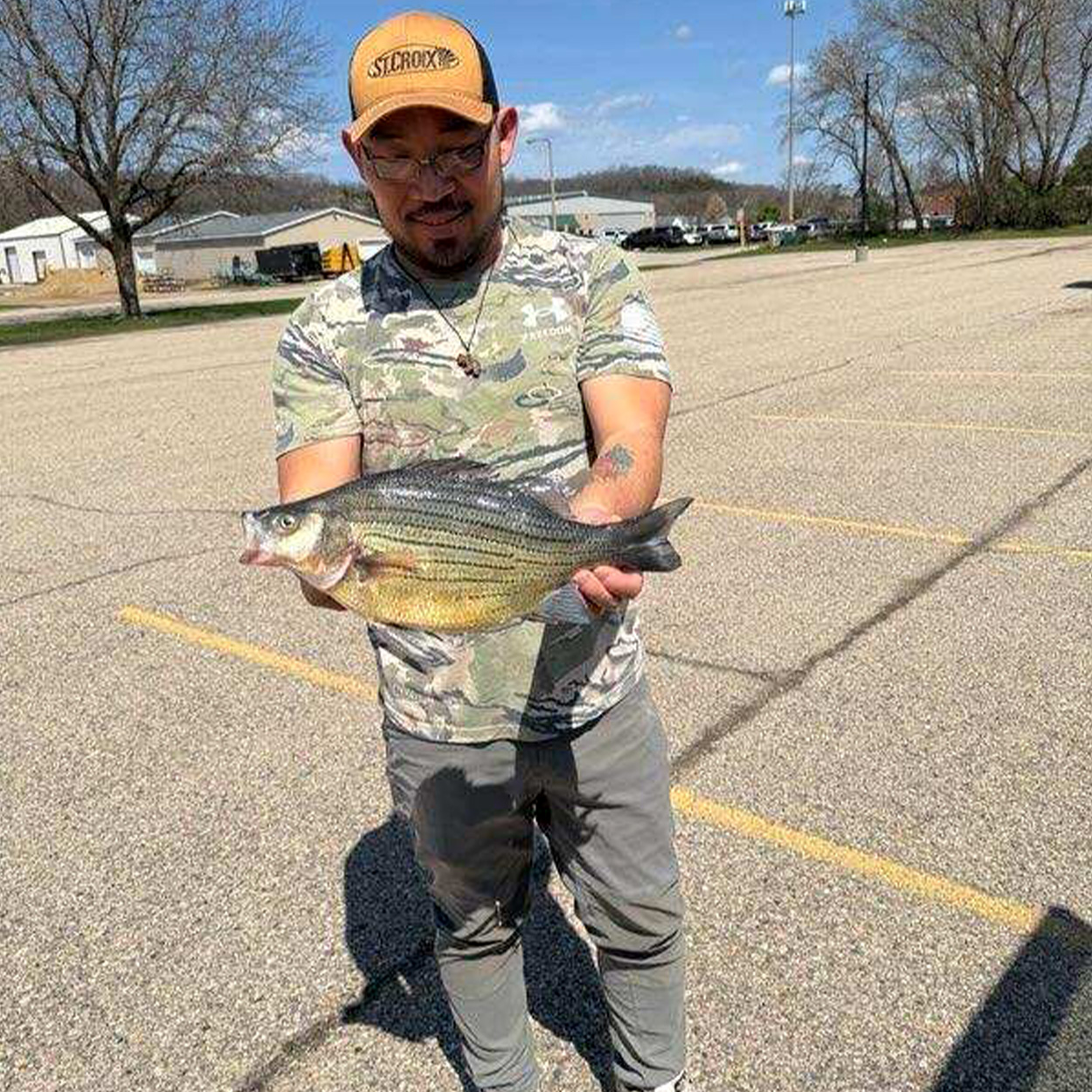 An angler holds up a yellow bass in a parking lot.