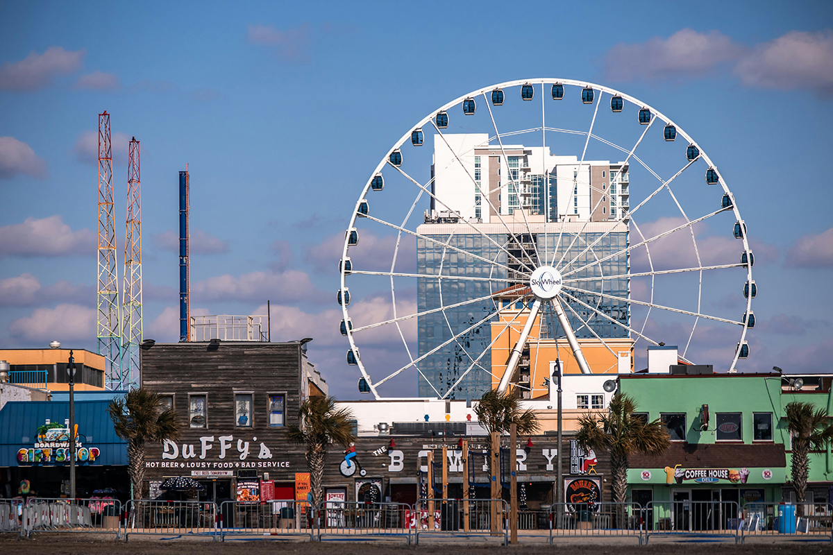 Ferris wheel towers over shops lining a boarwalk.