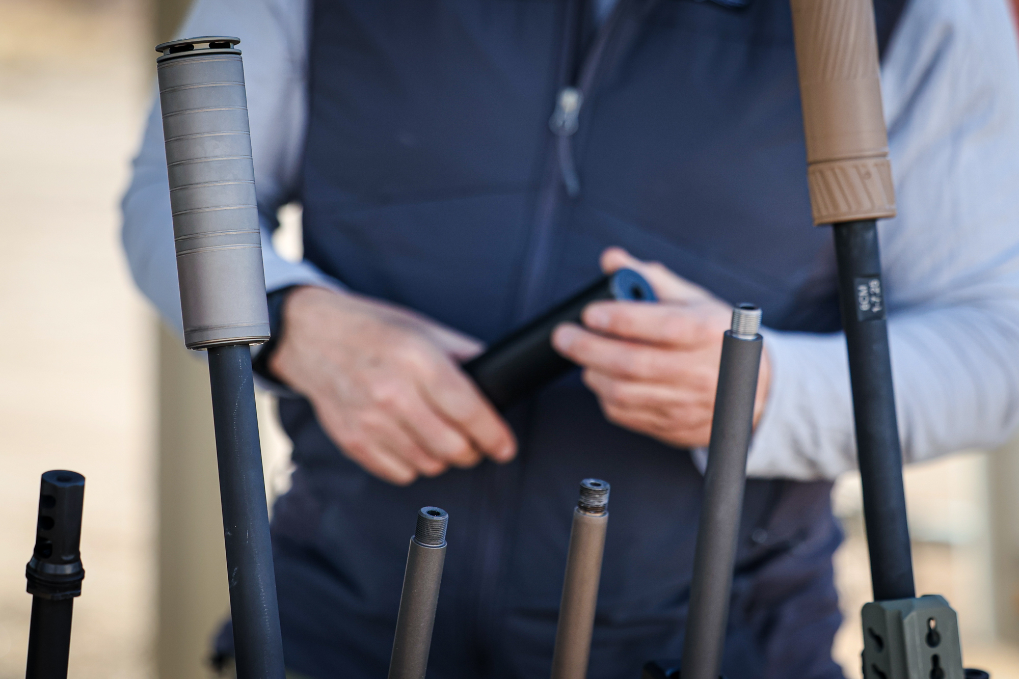 A man holds a suppressor by a rack of rifles, some with threaded barrels.