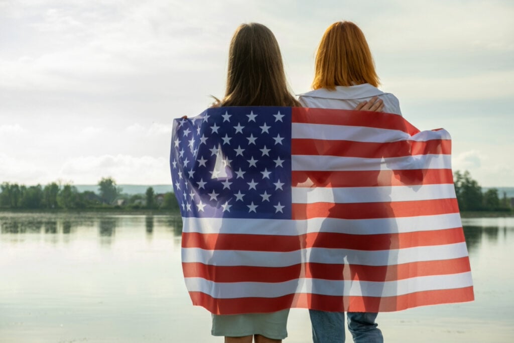 girls wrapped in American flag, looking at lake on Memorial Day