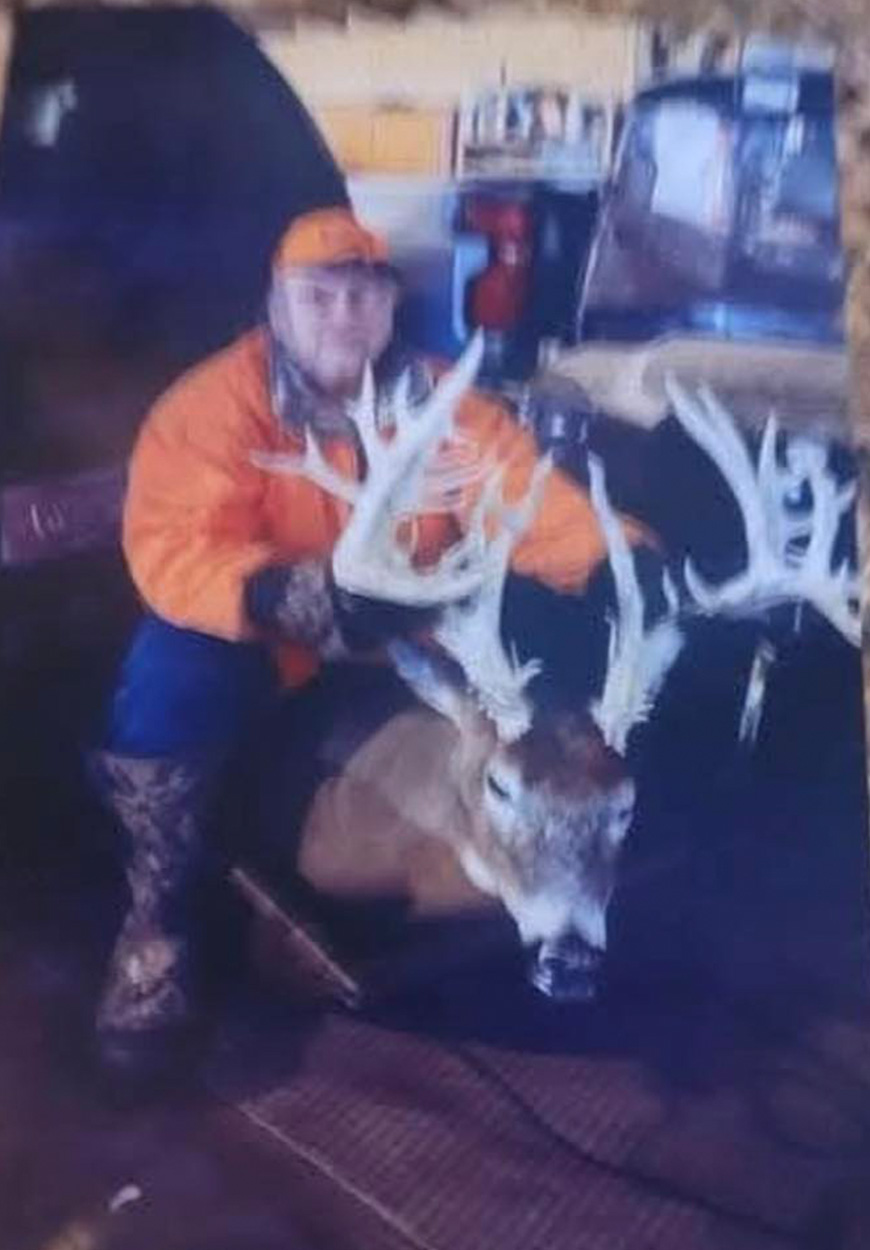 A deer hunter sits next to a deer behind a vehicle.
