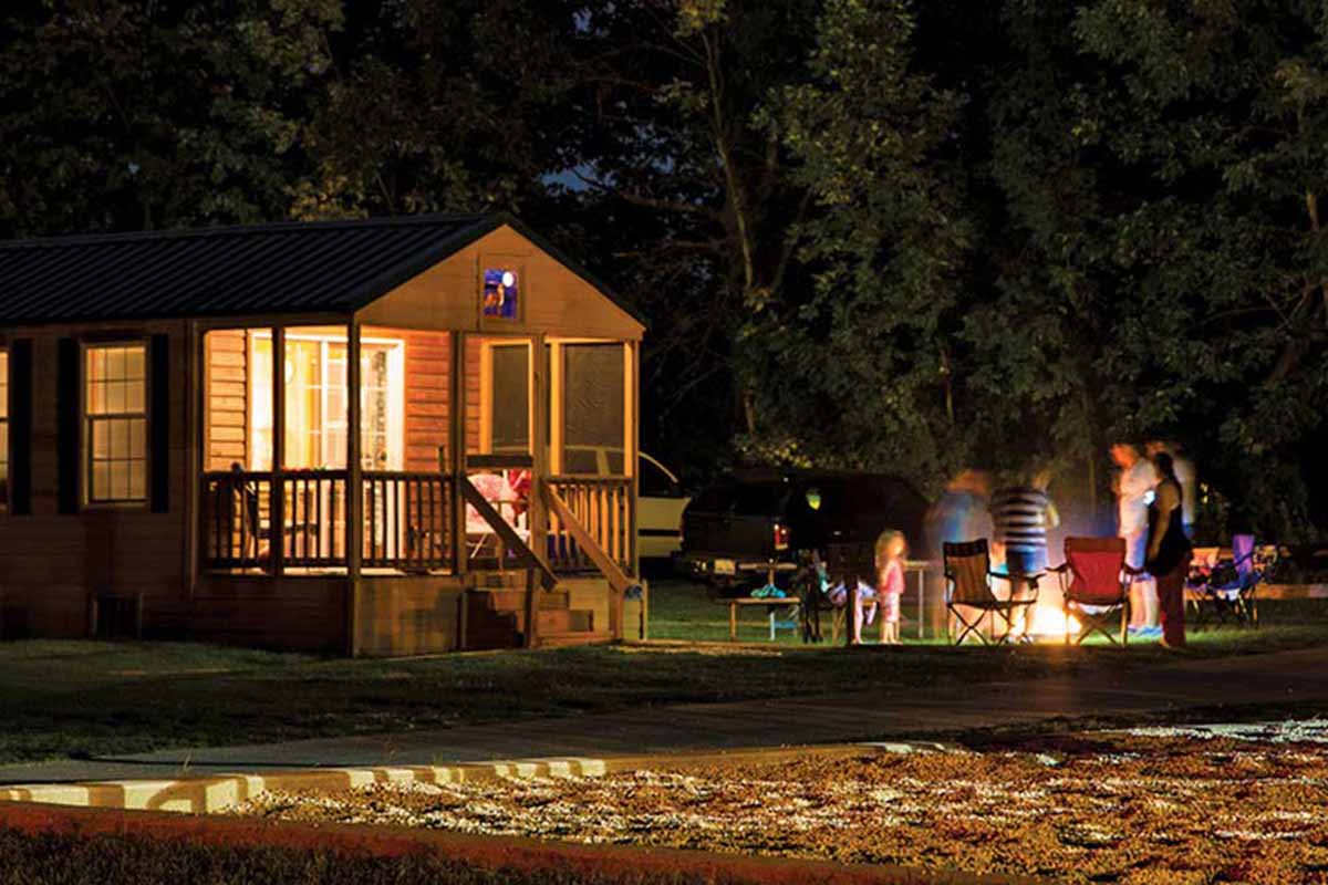 Cabin at night — nearby people roast marshmallows over fire.